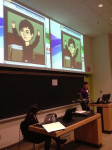 In a classroom, a white woman in a purple men's dress shirt and short hair reads from a laptop. Above her is projected, side-by-side, identical images of cartoon rendition of another white woman in a long-sleeved black shirt, with short black hair, her arms lifted up triumphantly.