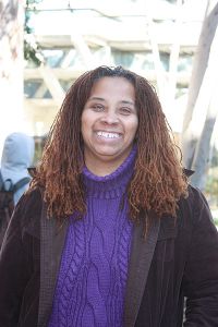 An African American woman, smiling, with shoulder-length hair, in a purple knitted turtleneck with a black jacket over it.